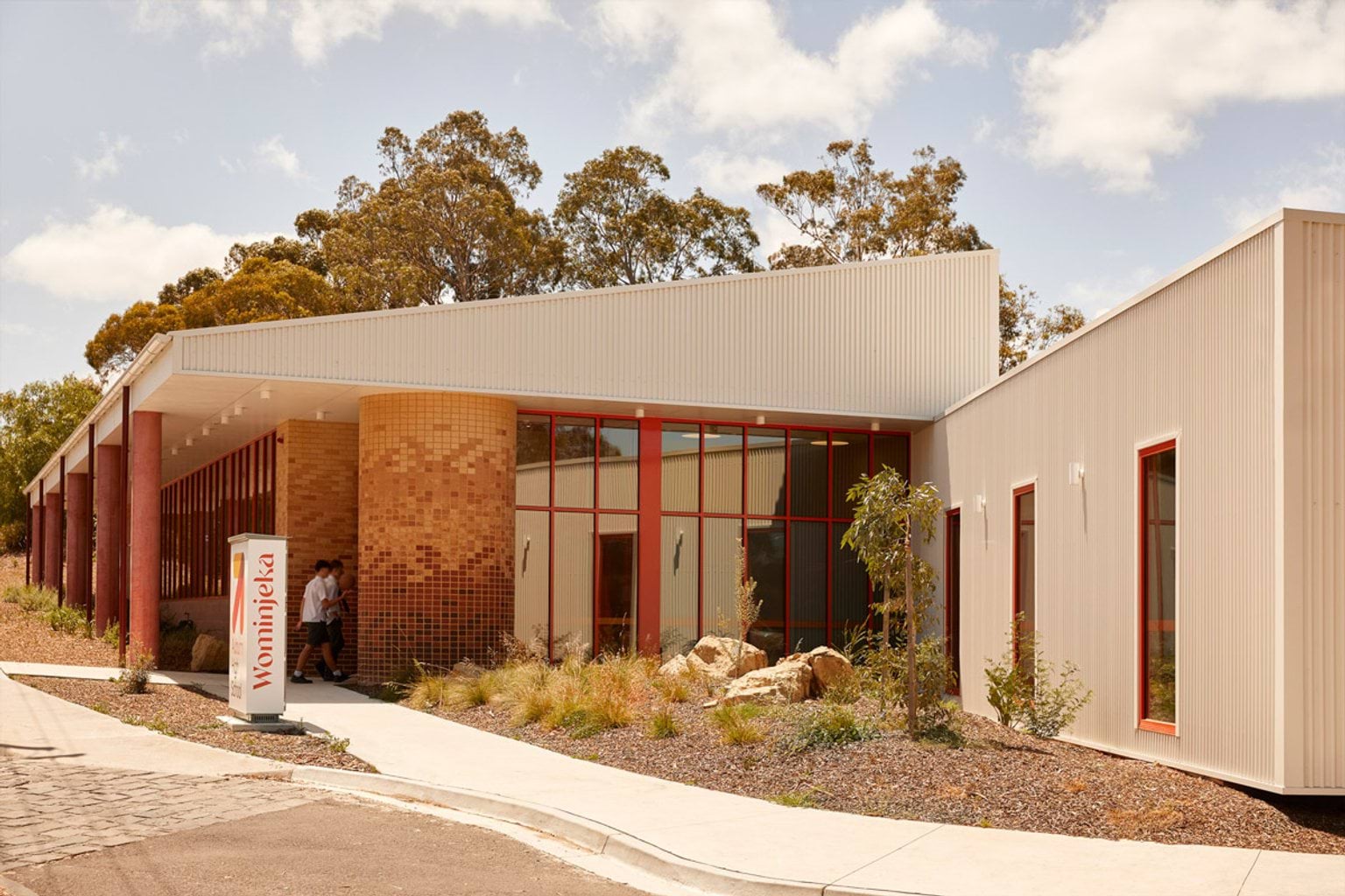 The main entrance of a school building with angled rooflines, brick columns, and red-framed windows. A landscaped garden with rocks and shrubs borders the walkway.