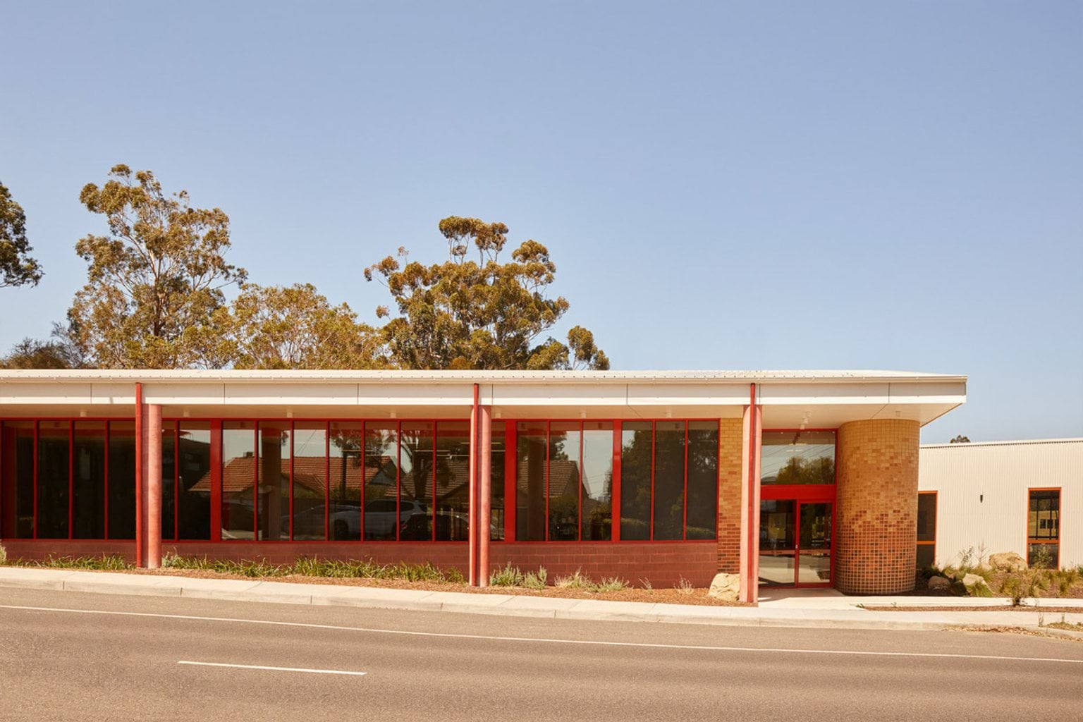 The exterior of a modern school building with large red-framed windows and brick columns, set against a clear blue sky with trees in the background.