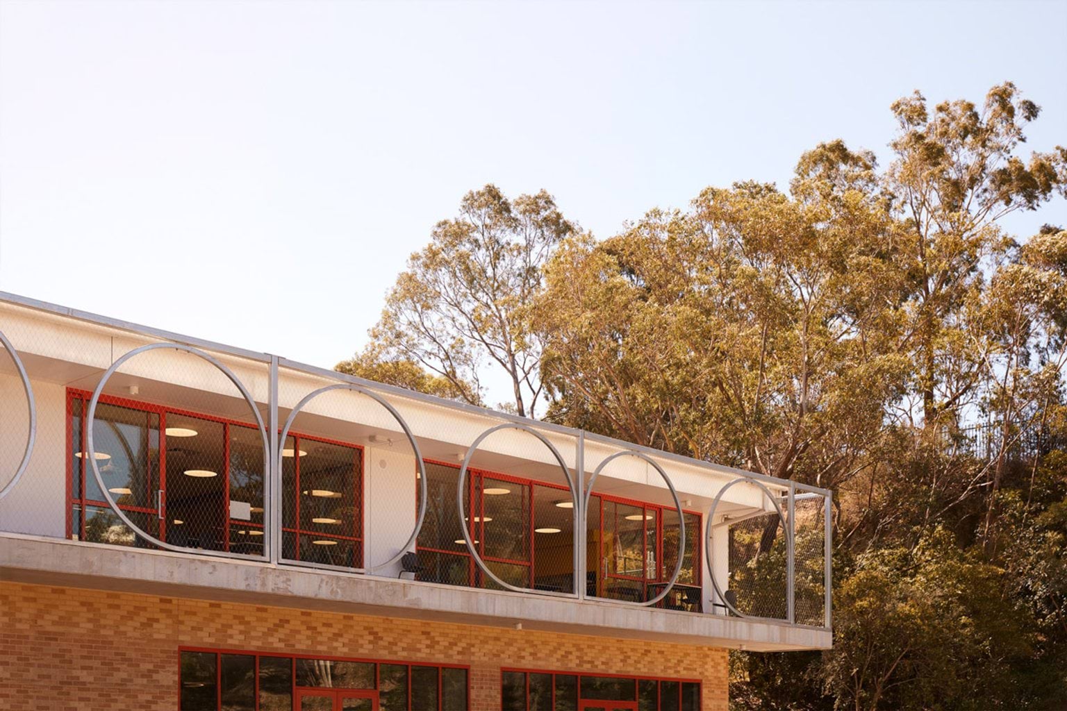 The upper level of a school building featuring a balcony enclosed by large circular metal frames and mesh. Red-framed windows line the building behind the balcony.