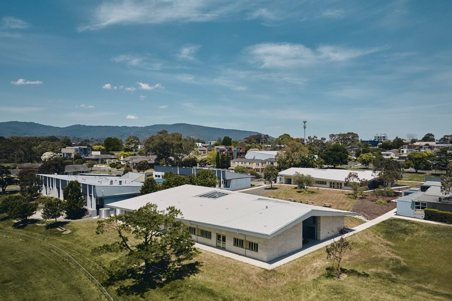 An aerial view of a modern school campus with multiple low-rise buildings, landscaped paths, and surrounding greenery, set against a backdrop of hills.