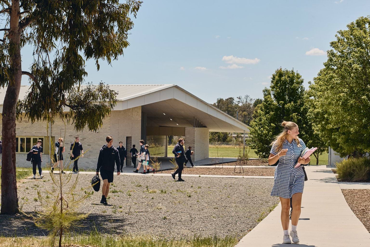 Students walking along a paved path outside a light brick school building with a wide roof and open entryway, surrounded by trees and garden beds.