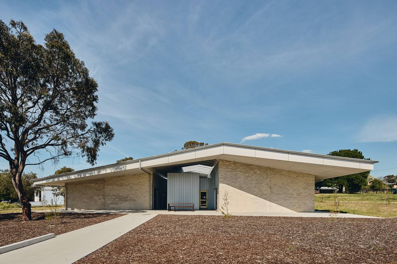 A single-storey school building with pale brick walls and a wide angled roof, set in a landscaped area with a concrete path and young trees.