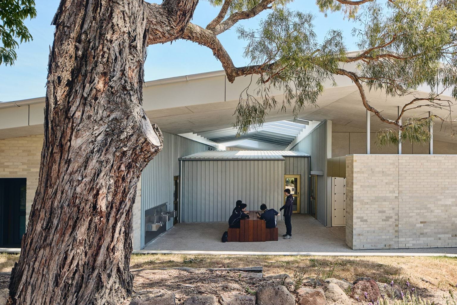 A shaded outdoor space with timber tables and benches under a slatted roof, framed by corrugated walls and landscaping.