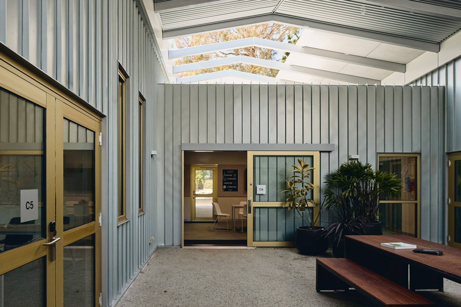 A courtyard with timber bench seating, potted plants, and yellow-framed doors under a slatted roof allowing natural light.