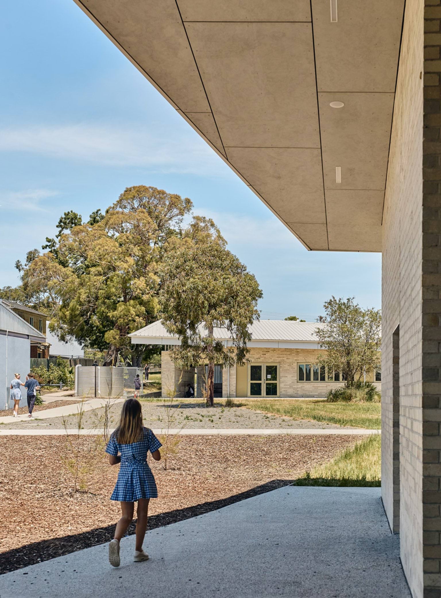 A student walking along a concrete path in a landscaped courtyard between modern school buildings with wide roofs and light brick walls.