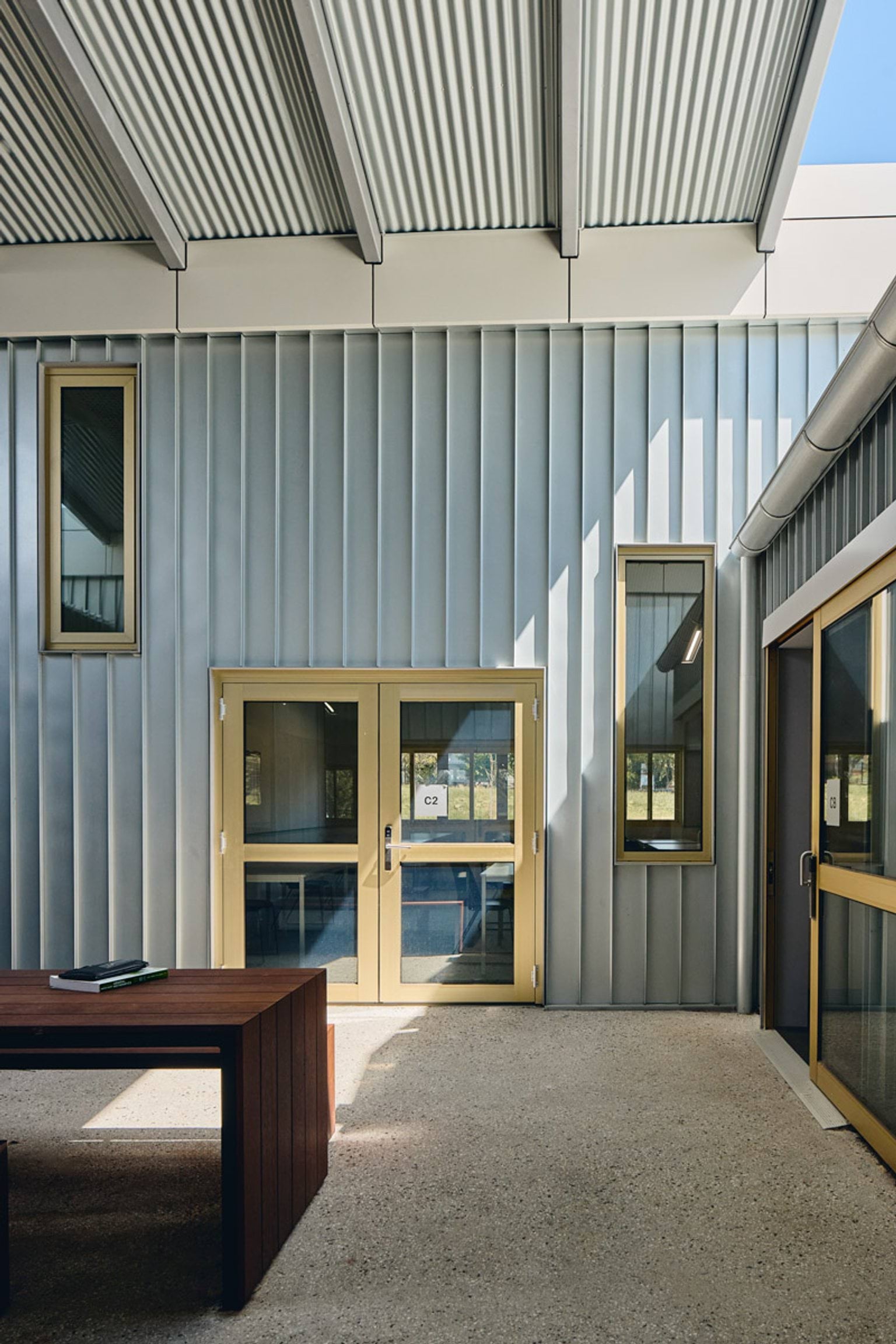 A covered outdoor area with corrugated metal walls, yellow-framed glass doors, and a timber table under a slatted roof.