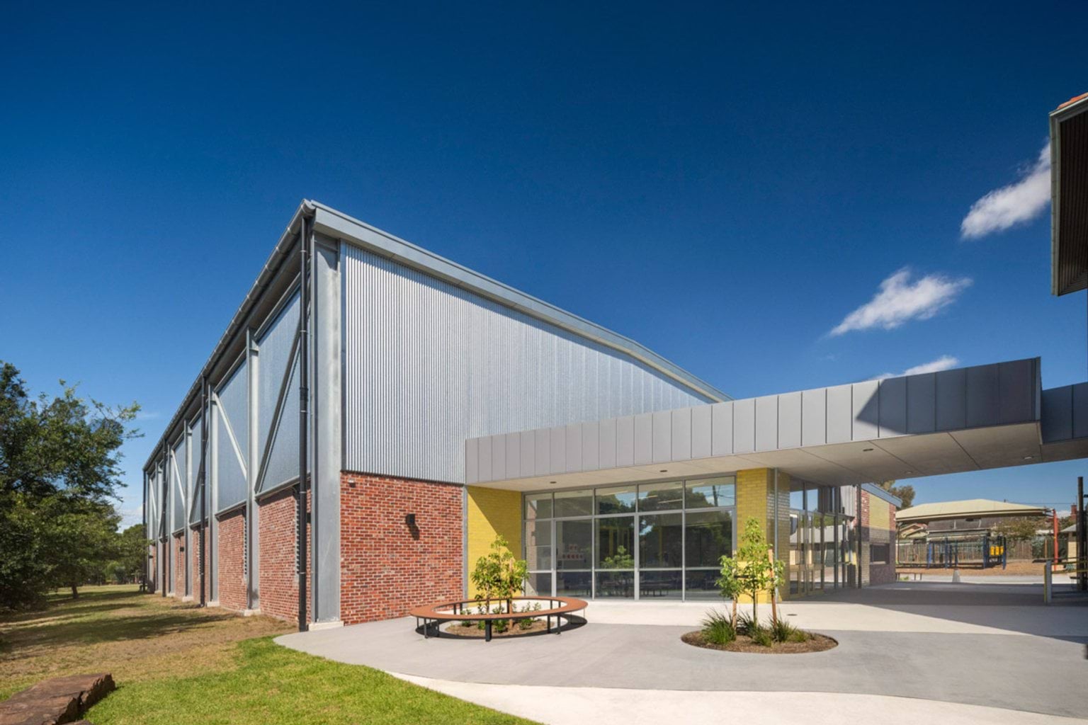 A modern school entrance with red brick and yellow feature walls, large glass windows, and a landscaped courtyard with a circular bench.