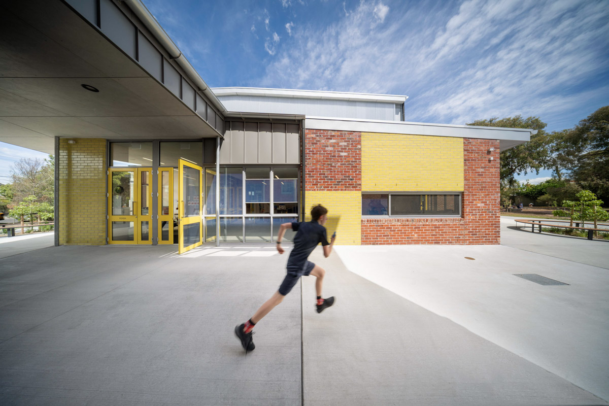 A student running across a paved area outside a modern school building with red brick walls, yellow feature panels, and glass windows.