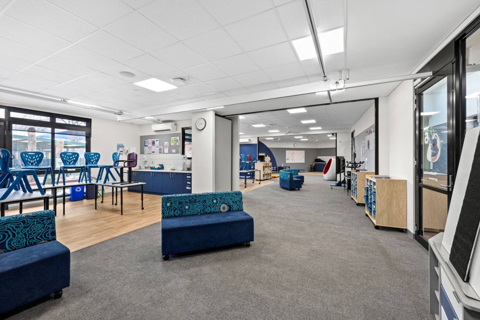 An open learning space with grey carpet, blue upholstered seating, and a kitchenette. Movable partitions open to adjoining rooms.