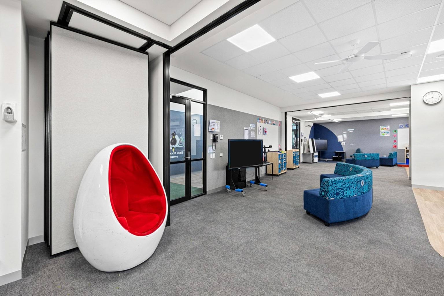An open learning space with grey carpet, blue seating, and a distinctive red egg-shaped chair. Movable partitions separate adjoining rooms.