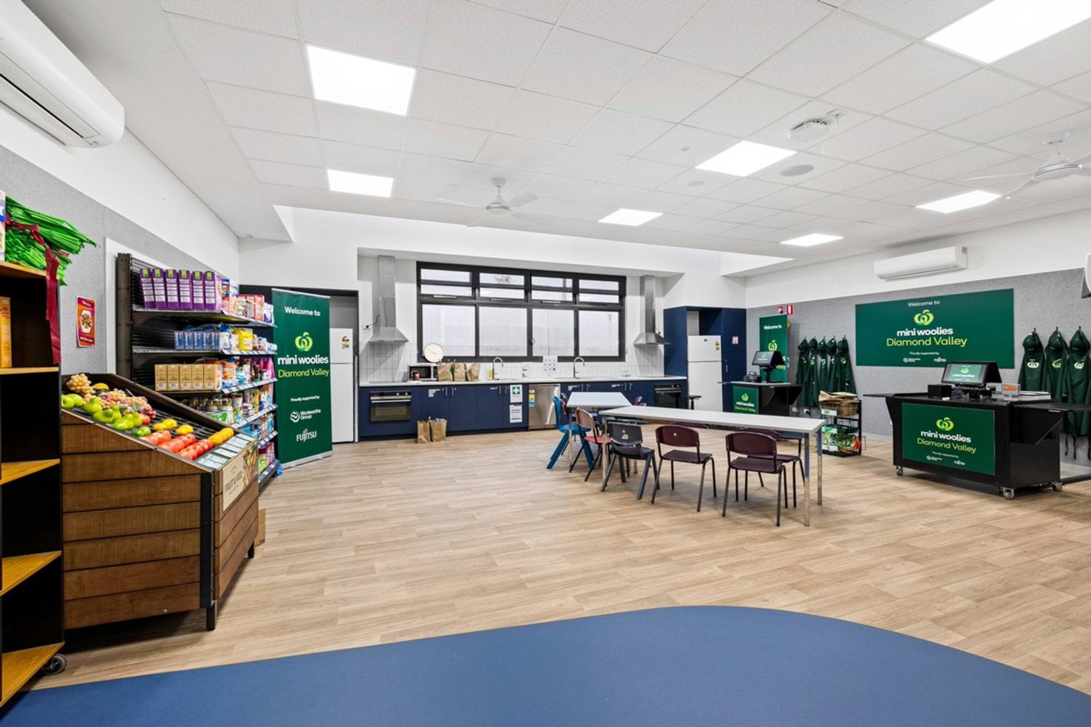 A classroom designed as a mini supermarket with shelves of groceries, green signage, and tables for activities. A kitchenette runs along the back wall.