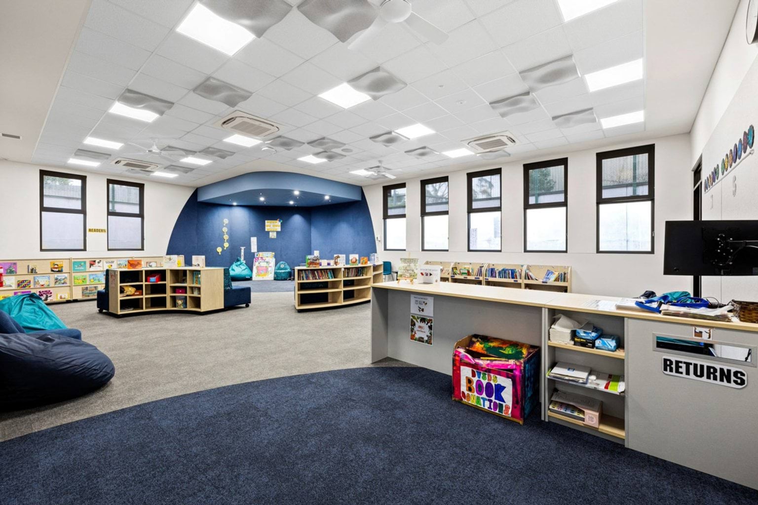A library with a returns desk, curved shelving units, beanbags, and a blue feature wall. Large windows provide natural light.