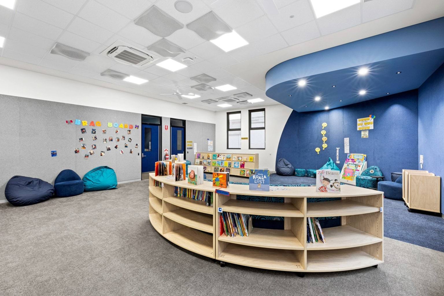 A library with grey carpet, curved blue seating, beanbags, and timber shelving filled with books. A blue feature wall with spotlights is in the background.