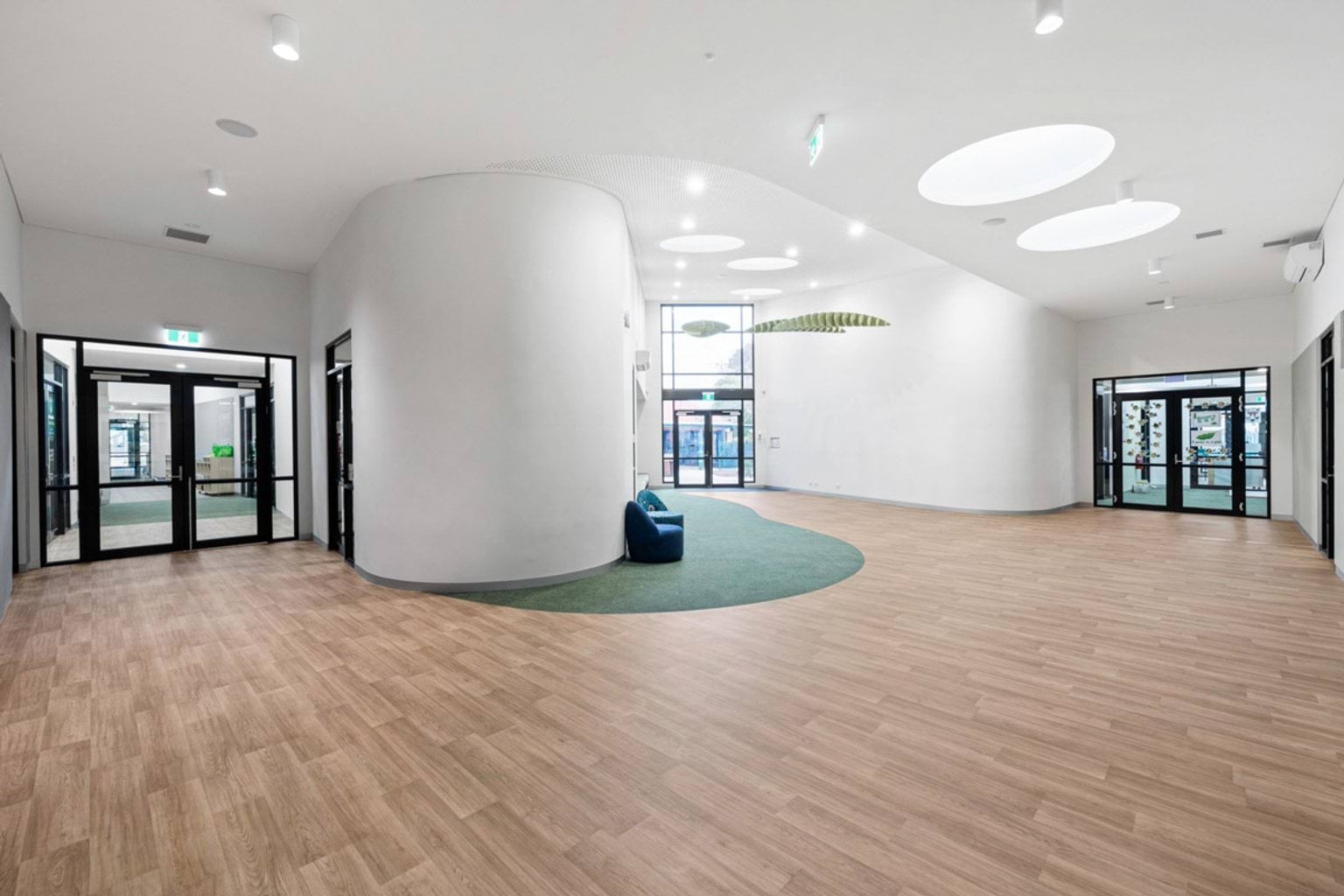 A spacious foyer with timber flooring, curved white walls, and circular ceiling lights. Green carpeted seating areas are integrated into the design.