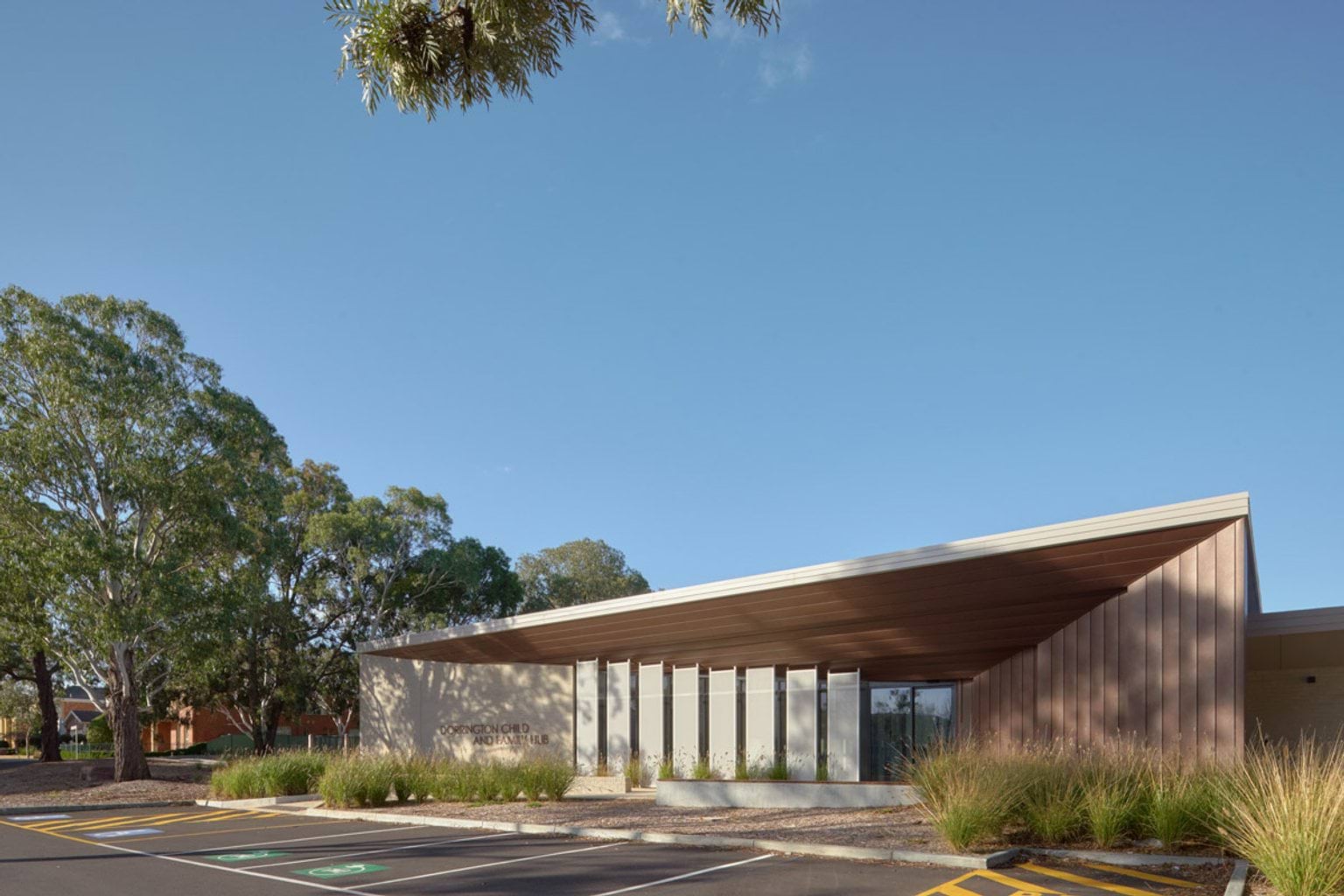 The exterior of the kindergarten with a wide timber-clad roof and angled facade panels. There are tall vertical screens, landscaped garden beds and a car park in the foreground with large gum trees behind.