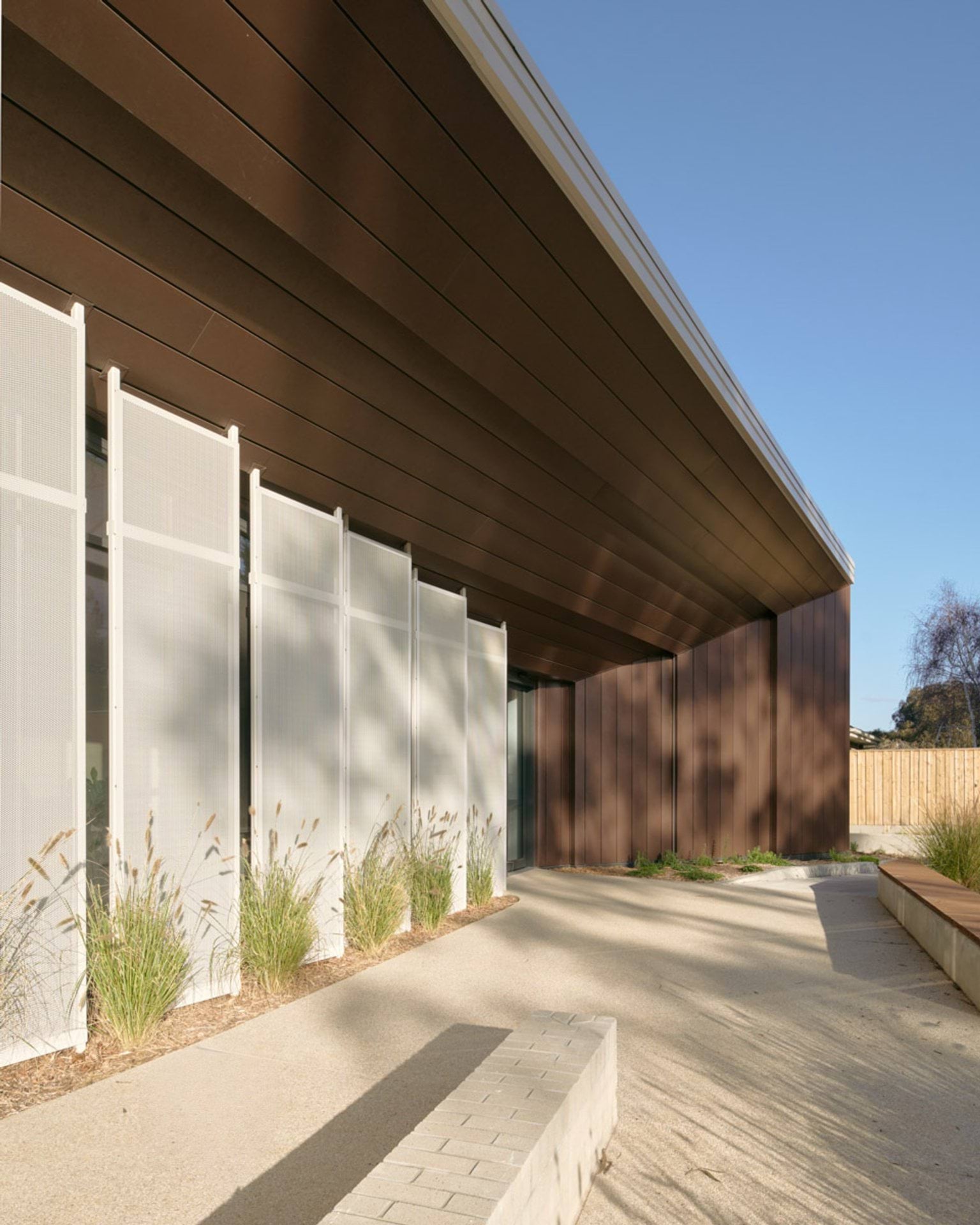 A close-up of the kindergarten entrance with angled timber panels and white vertical screens. There are landscaped garden beds with tall grasses and a paved path leading to the door.