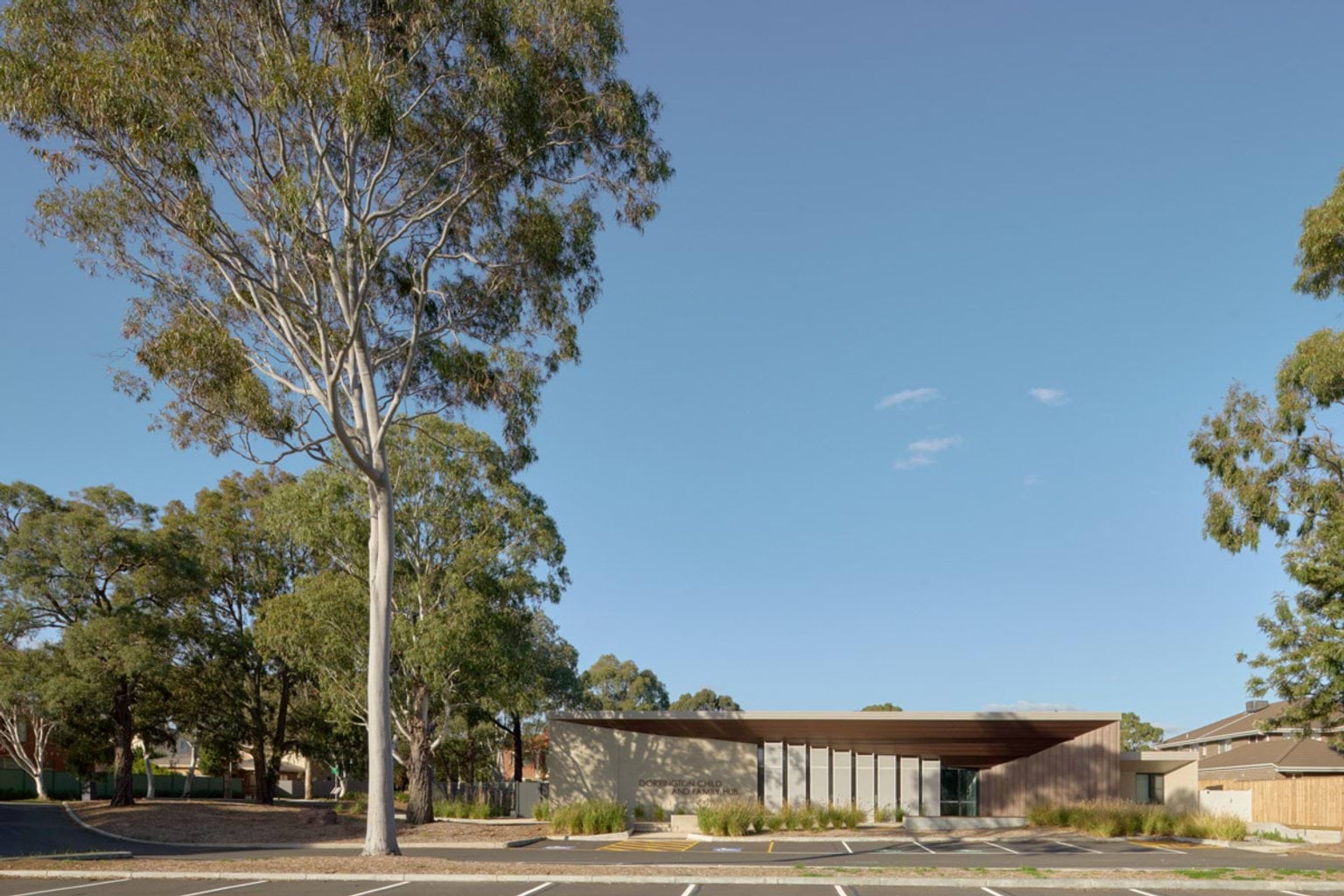 The exterior of the kindergarten viewed from the car park, with a wide timber-clad roof and vertical screens. Large gum trees surround the building under a clear blue sky.