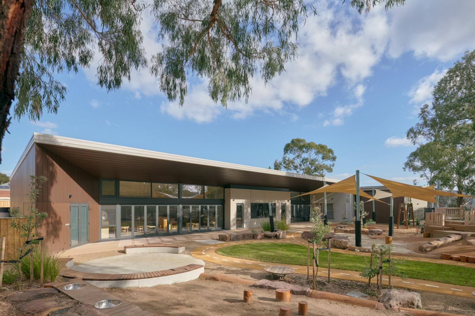 The exterior of the kindergarten viewed from the outdoor play area, with timber cladding and large windows. Shade sails cover part of the garden, which has curved paths, rocks and young trees.