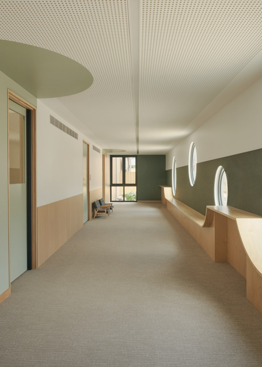 A hallway inside the kindergarten with round windows set into a green wall and timber bench seating below. The ceiling is white with perforated panels and the floor is carpeted in a neutral tone.