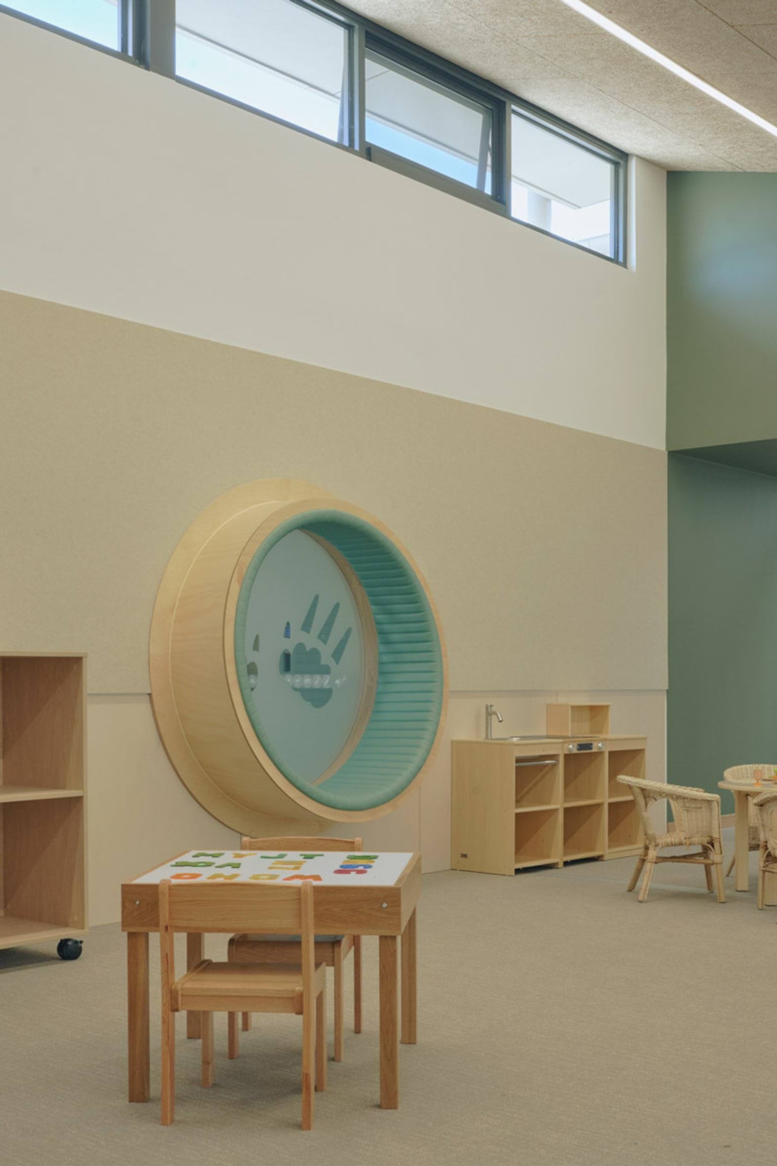 A kindergarten classroom with a round timber feature window and a small table with alphabet letters. There are timber shelves and wicker chairs against pale green walls.