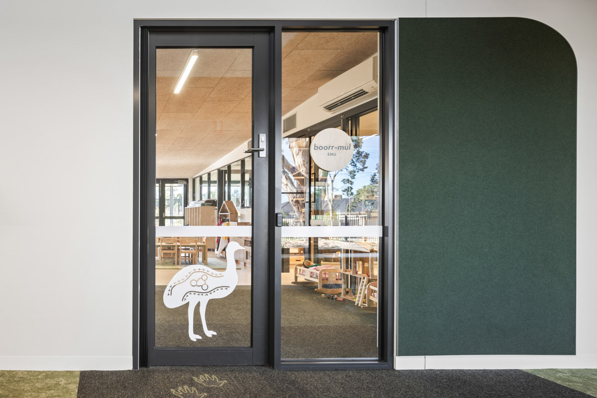 Classroom entrance with glass door displaying emu artwork and text, view into room with timber furniture and play equipment