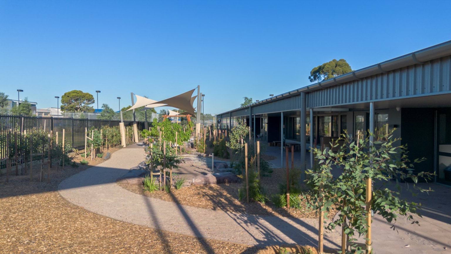 Outdoor play area with curved pathways, young plants and shade sails beside a modern single-storey building