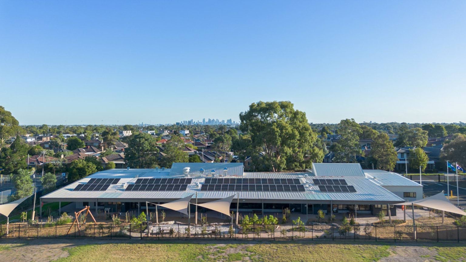 Aerial view of a single-storey building with solar panels on the roof, shade sails and surrounding residential area