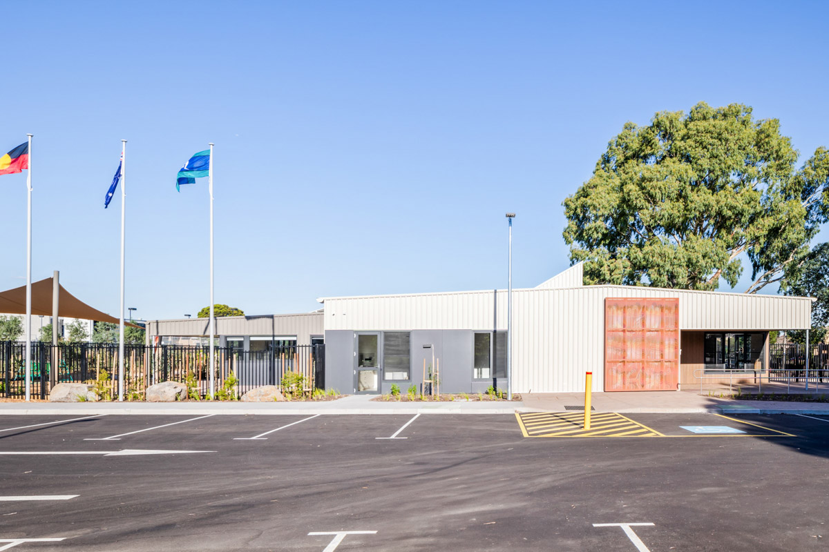 Front view of a modern building with metal cladding, large timber entry doors and three flagpoles in landscaped forecourt