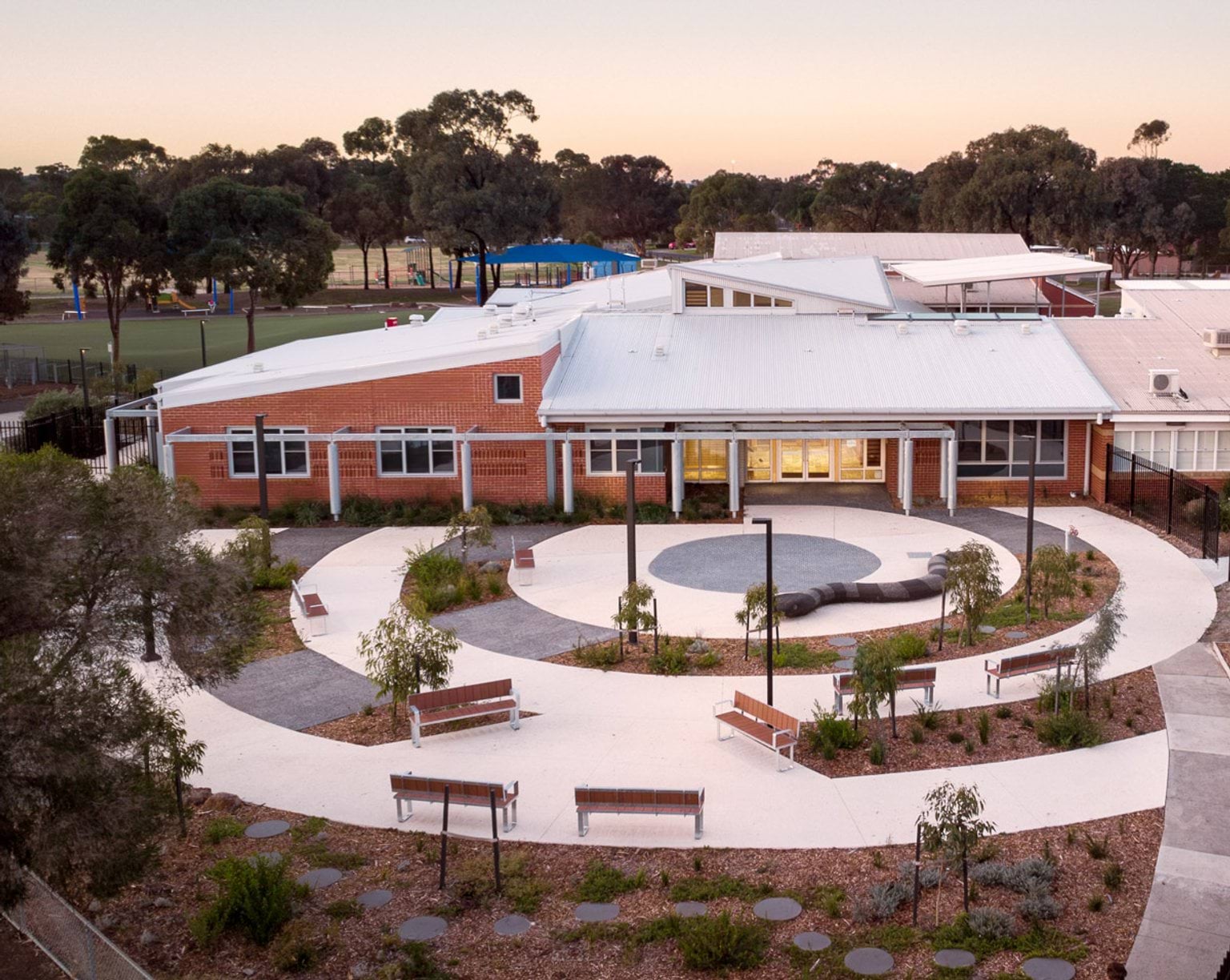 An aerial view of the school courtyard with curved paths, benches, and landscaped garden beds. The building has red brick walls and a white roof.