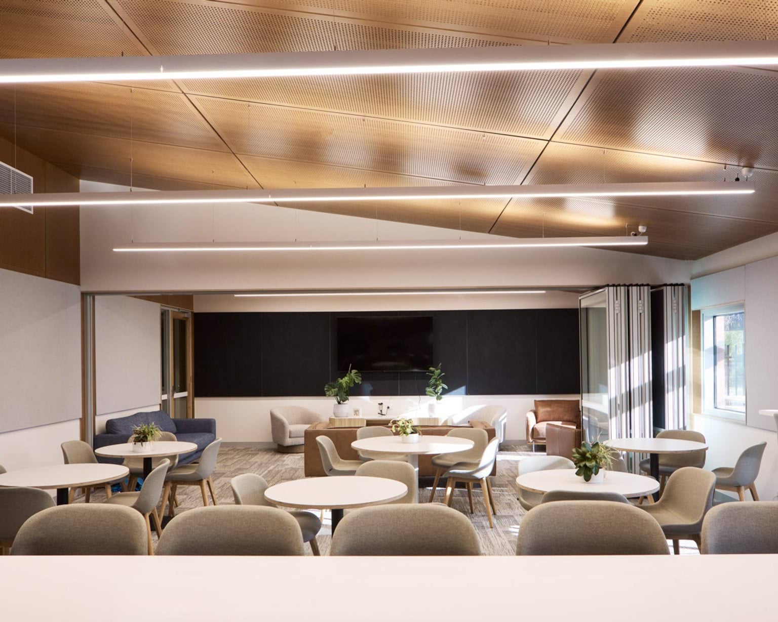 An indoor lounge with round tables and cushioned chairs. The space has a timber ceiling with linear lights and a large screen on the far wall.