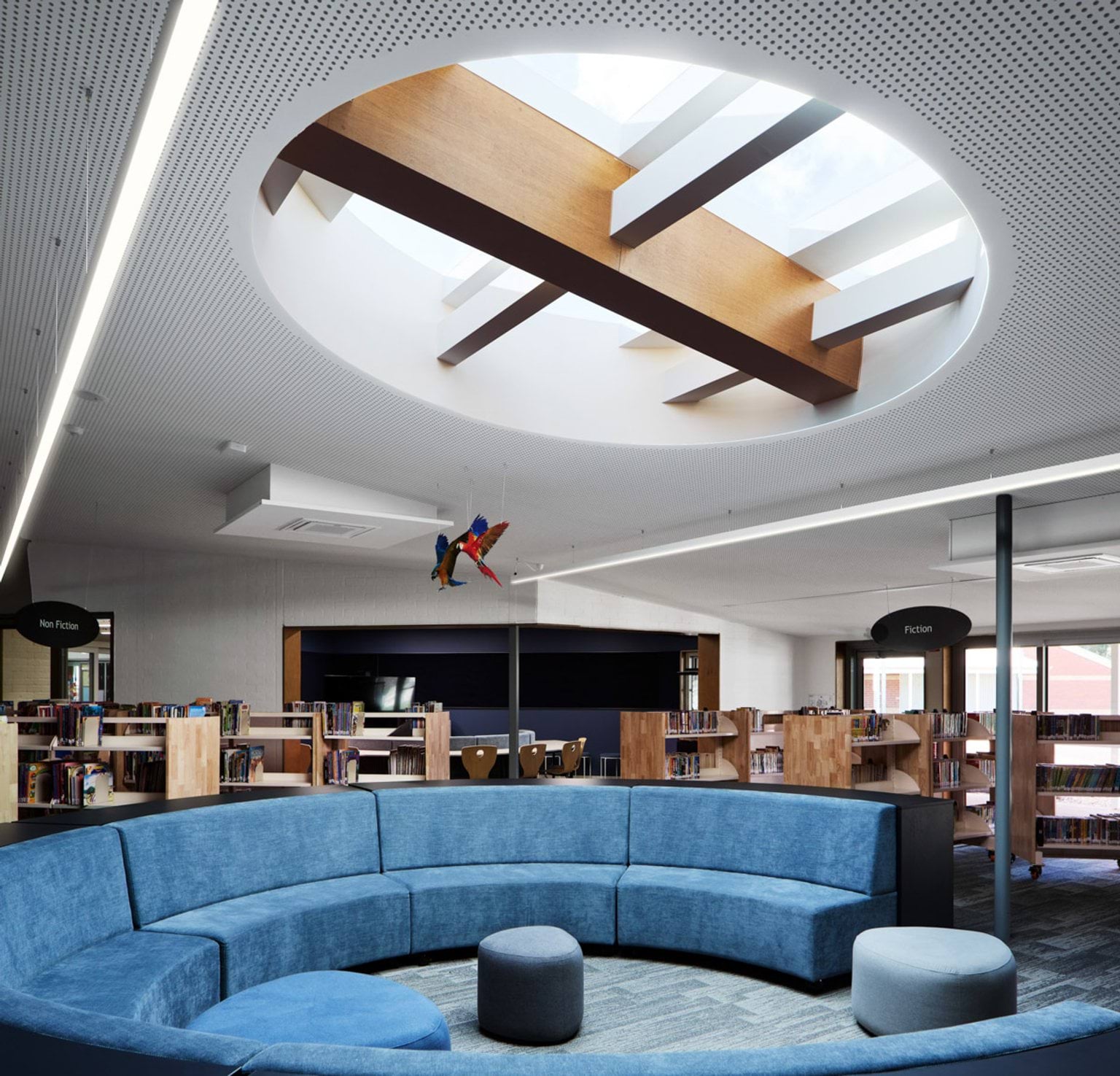 A library with a circular skylight and a blue curved lounge in the centre. Bookshelves line the walls beneath bright lighting.