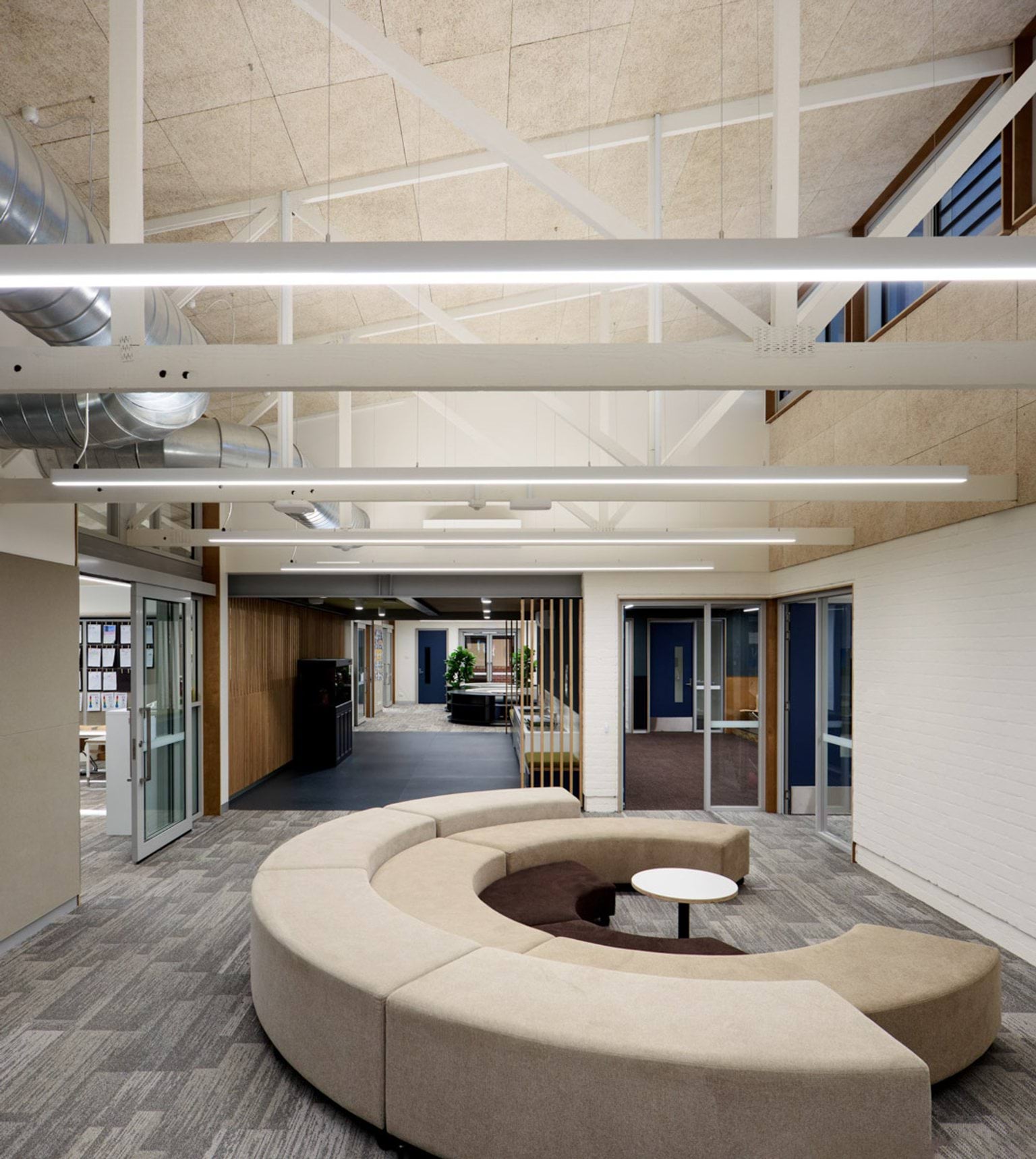 A bright foyer with high ceilings, exposed beams, and modern lighting. There is a curved beige lounge in the centre and glass doors leading to other rooms.