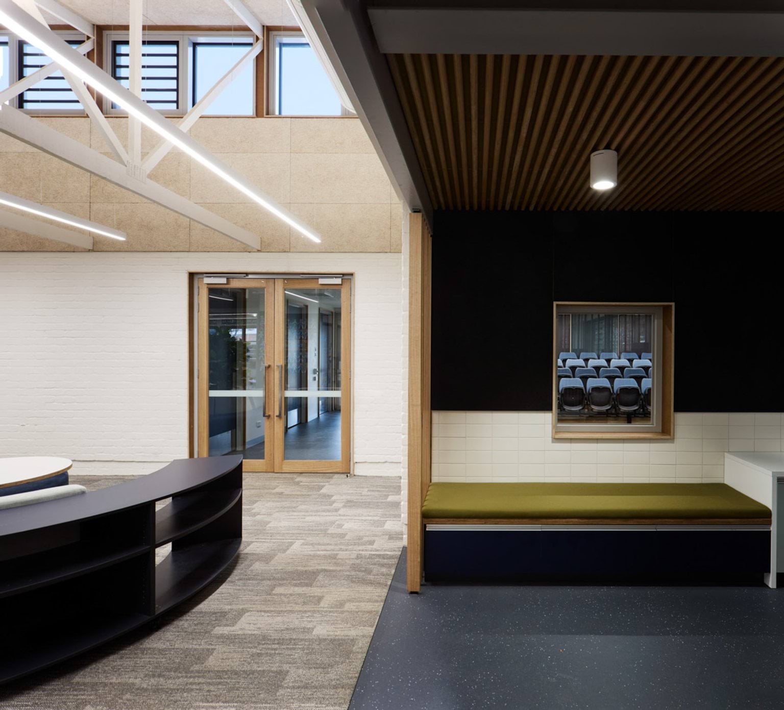 A hallway with timber accents and a built-in seating nook with a green cushion. Glass doors lead to another area of the school.