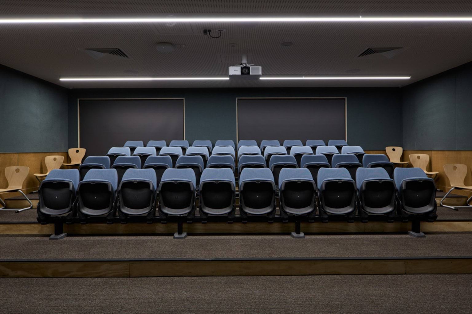 A small lecture room with tiered seating in rows facing a projector screen. The walls are dark with timber accents.