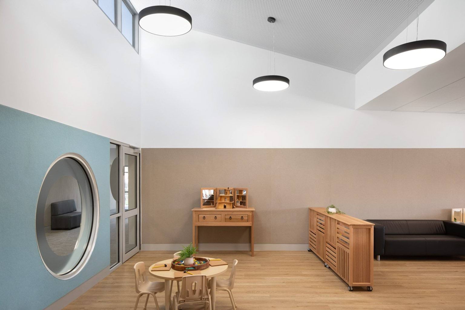 A kindergarten classroom with a round window set into a blue wall, timber furniture and a black couch. There is a small round table with chairs and craft materials, and large circular ceiling lights above.