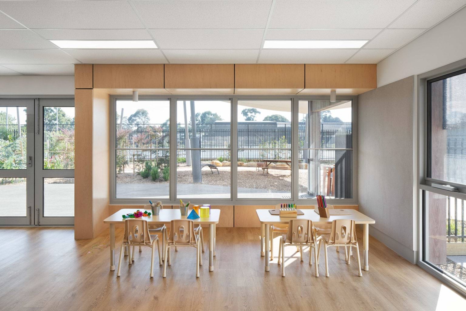 A bright kindergarten classroom with timber tables and chairs arranged near large windows. Colourful craft materials and pencils are on the tables, and the outdoor play area is visible through the glass doors.