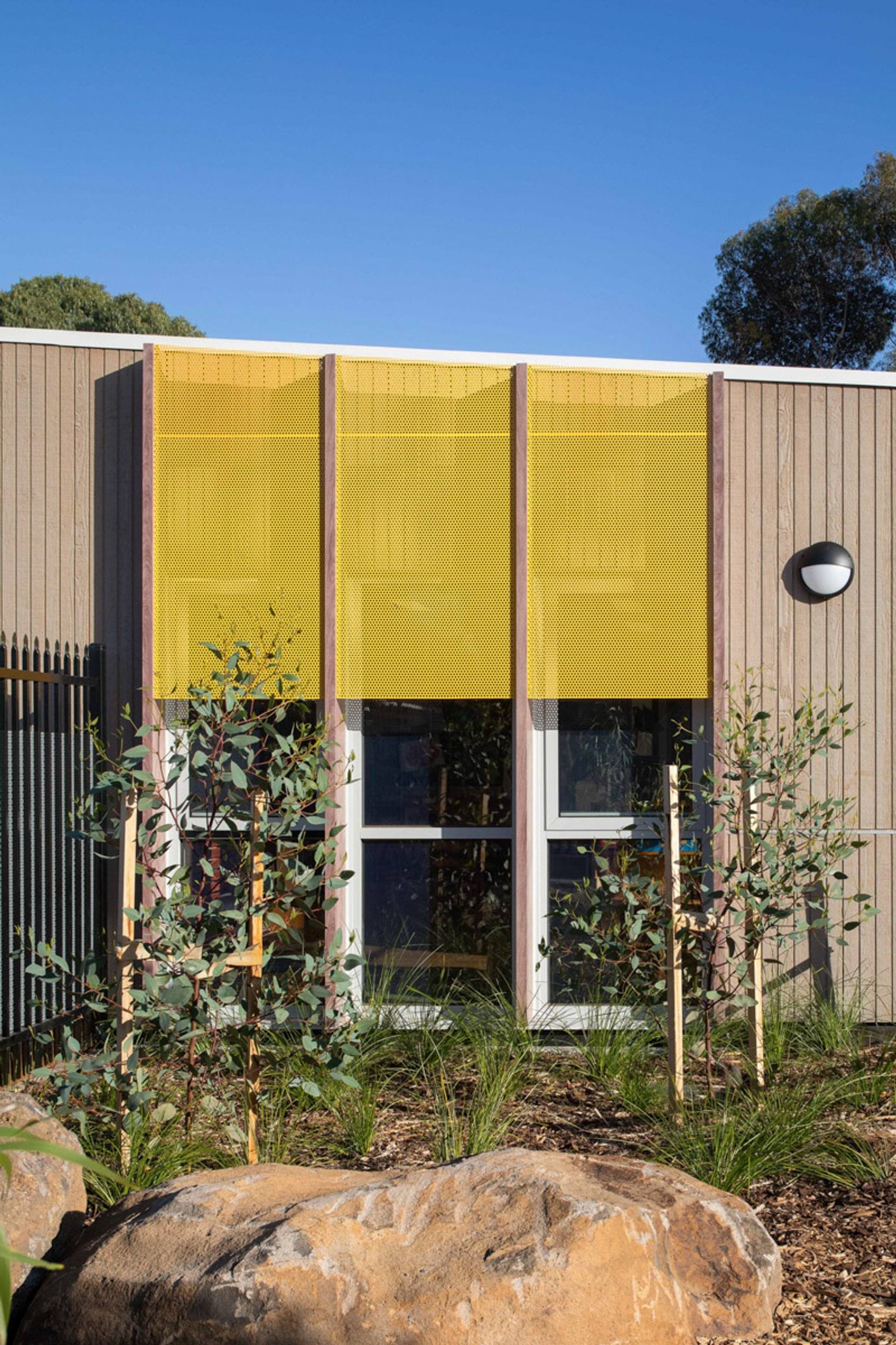 The exterior of the kindergarten with vertical timber panels and bright yellow perforated screens above the windows. There are young native plants and large rocks in the landscaped garden bed.