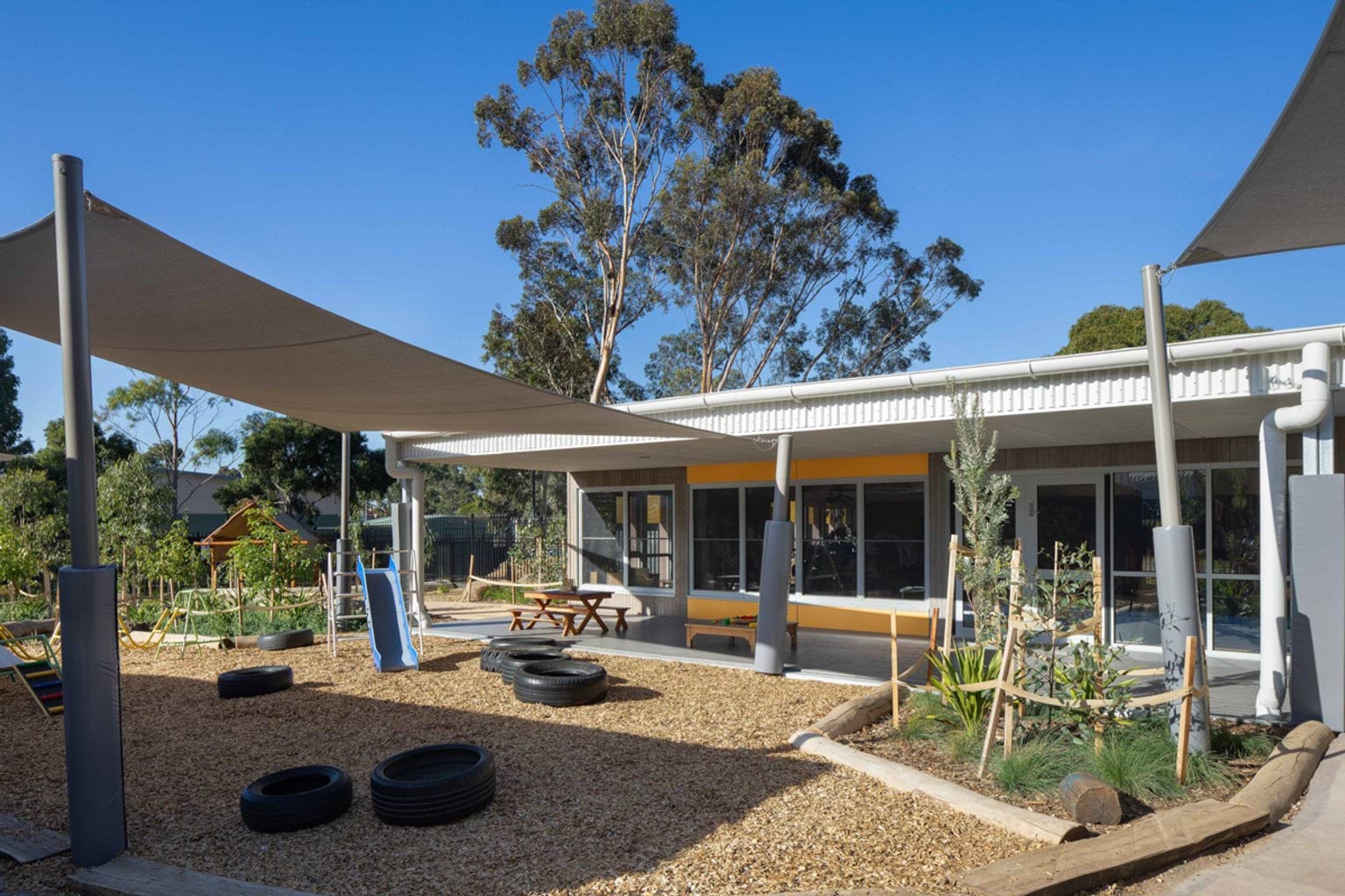 The outdoor play area of the kindergarten with shade sails, a blue slide and timber play equipment. There are tyres on a mulch surface and garden beds with young plants around the edge.
