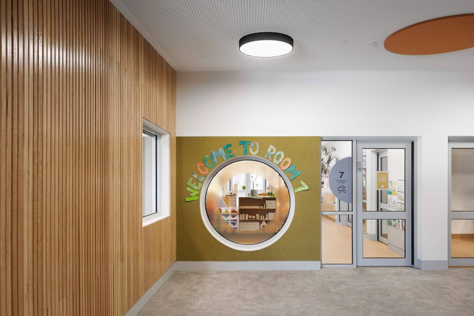 A hallway inside the kindergarten with timber wall panels and a circular window framed by green felt. Above the window, colourful letters spell “Welcome to Room 7”, and the classroom can be seen through the glass doors.