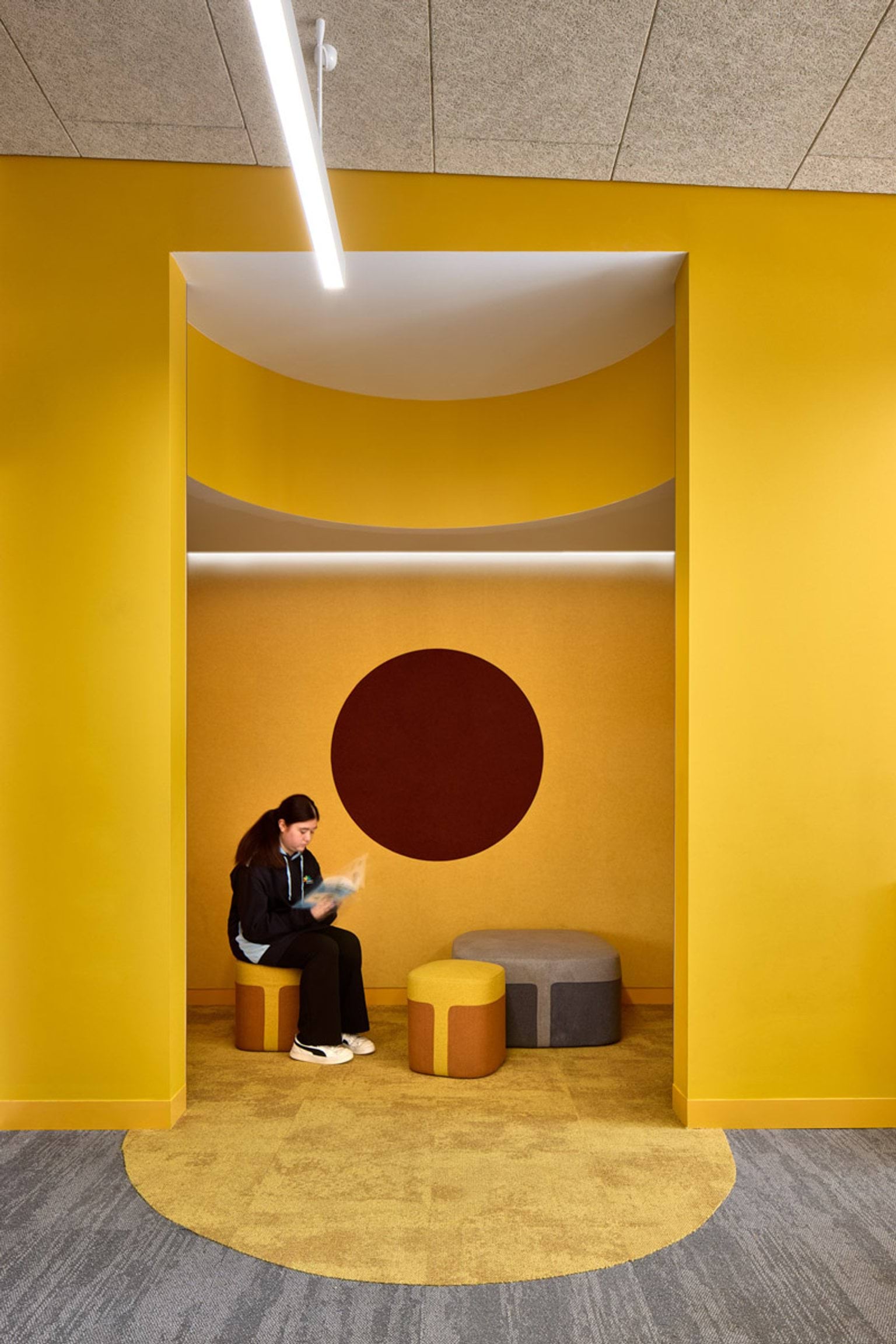 A cozy reading nook with bright yellow walls and a circular design feature. Small cushioned stools are arranged under a linear light.