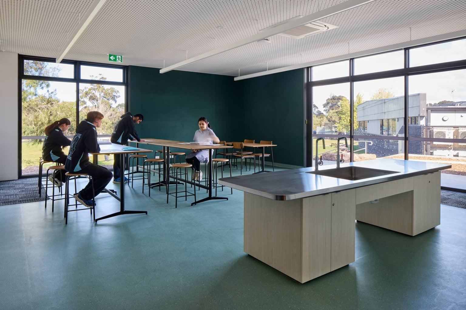 A classroom with green flooring, large windows, and high benches arranged in rows. A central island bench with a sink is in the foreground.