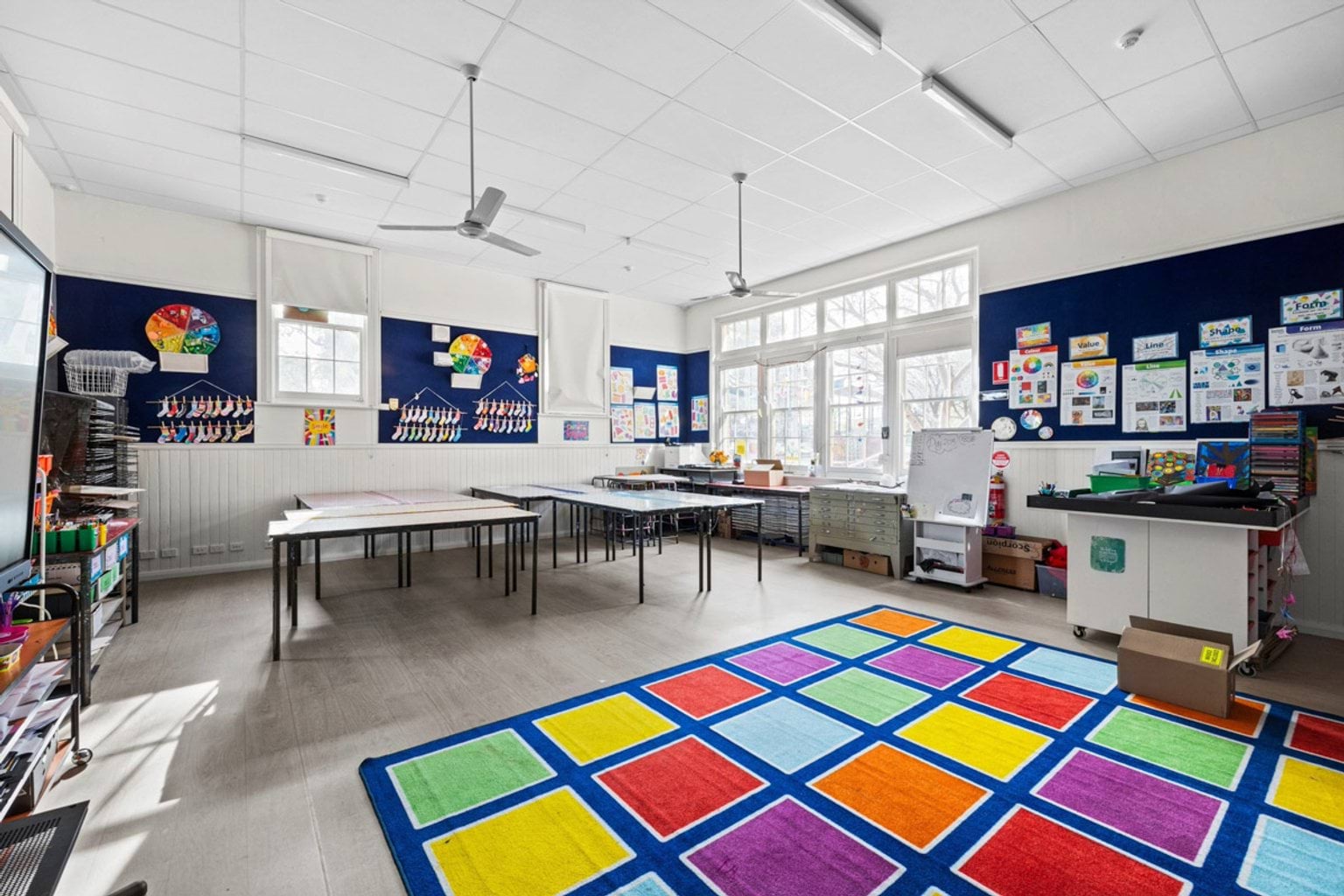 A bright classroom with large windows, white walls, and a colourful square-patterned rug. Tables and chairs are arranged for group activities, and student artwork decorates the walls.
