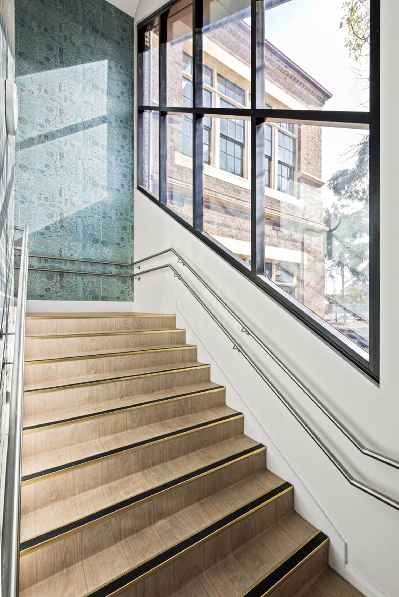 A staircase with timber steps and metal handrails next to a large window overlooking the heritage school building outside.