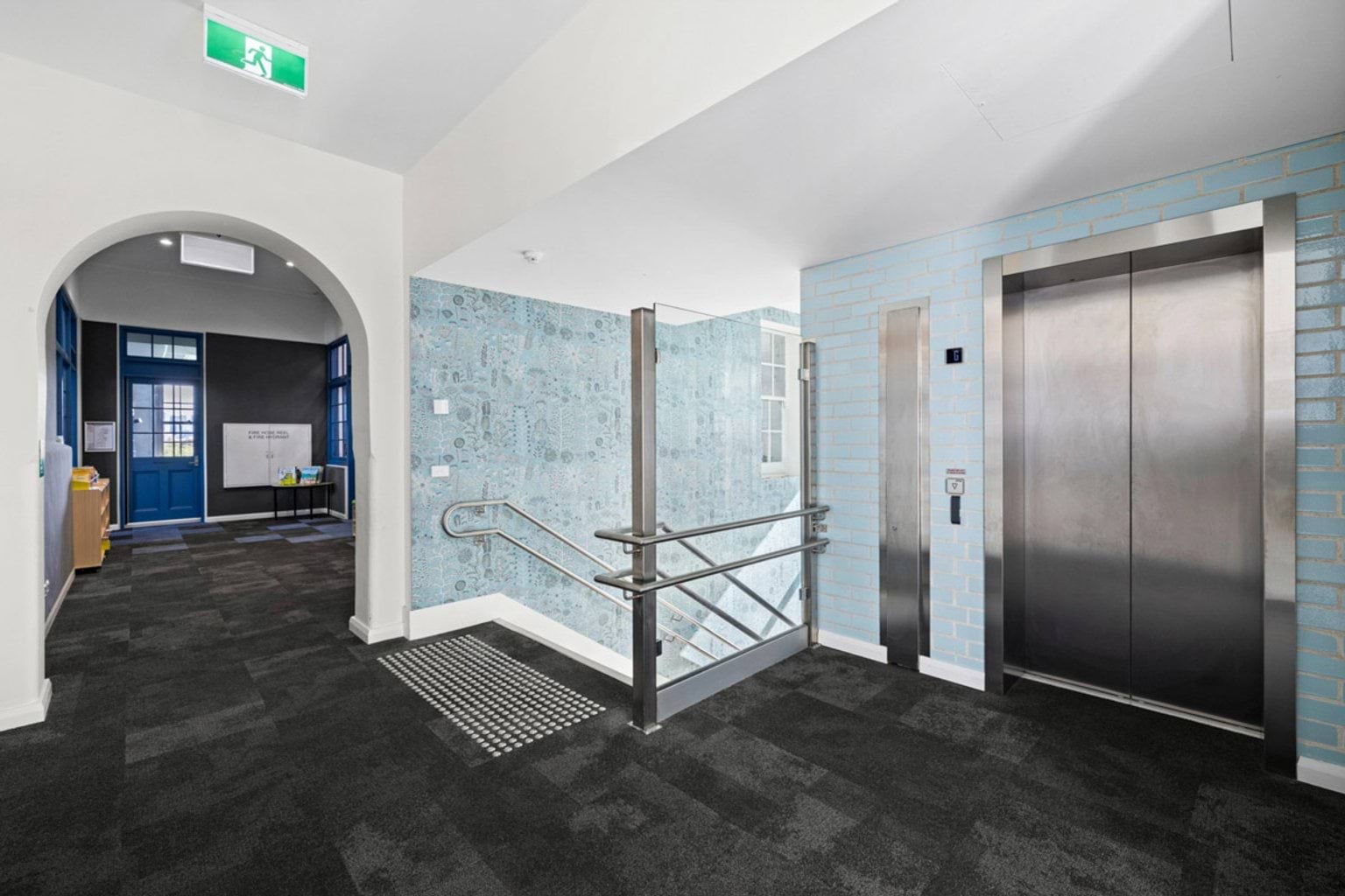 A lift lobby with a stainless steel lift, blue tiled walls, and black carpet flooring. An arched doorway leads to another corridor.