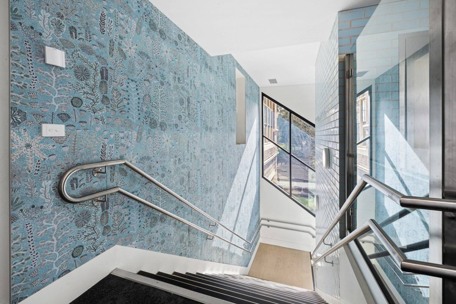 A staircase with metal handrails and a patterned blue feature wall, leading down to a landing with a large glass window.