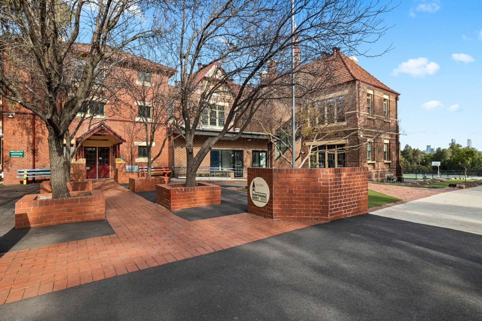 The exterior of a heritage red brick school building with pitched roofs and a paved courtyard featuring garden beds and trees.