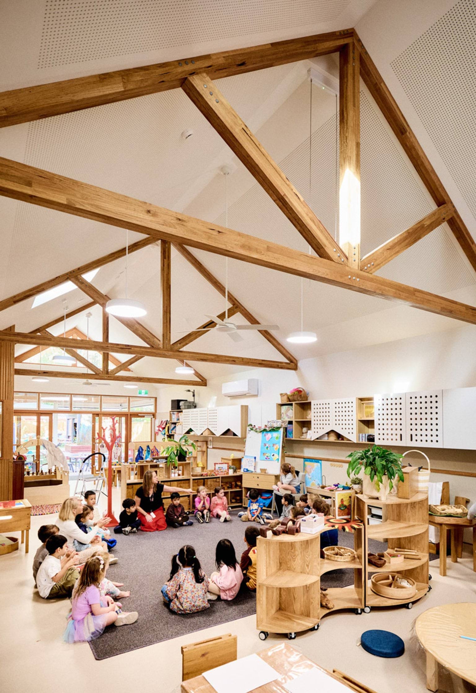 A kindergarten classroom with exposed timber trusses and pendant lights. Children are seated in a circle on a grey rug, surrounded by timber shelves and colourful artwork.