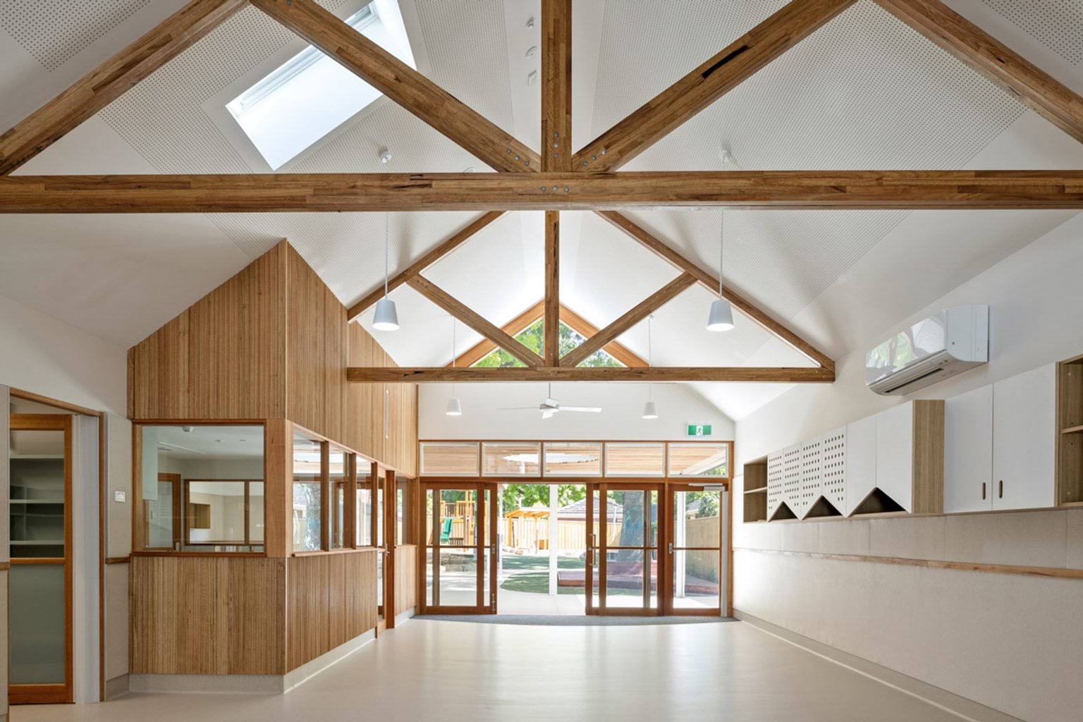 The interior of the kindergarten with a pitched ceiling and exposed timber trusses. Timber-framed glass doors open to the outdoor area, and built-in storage runs along one wall.