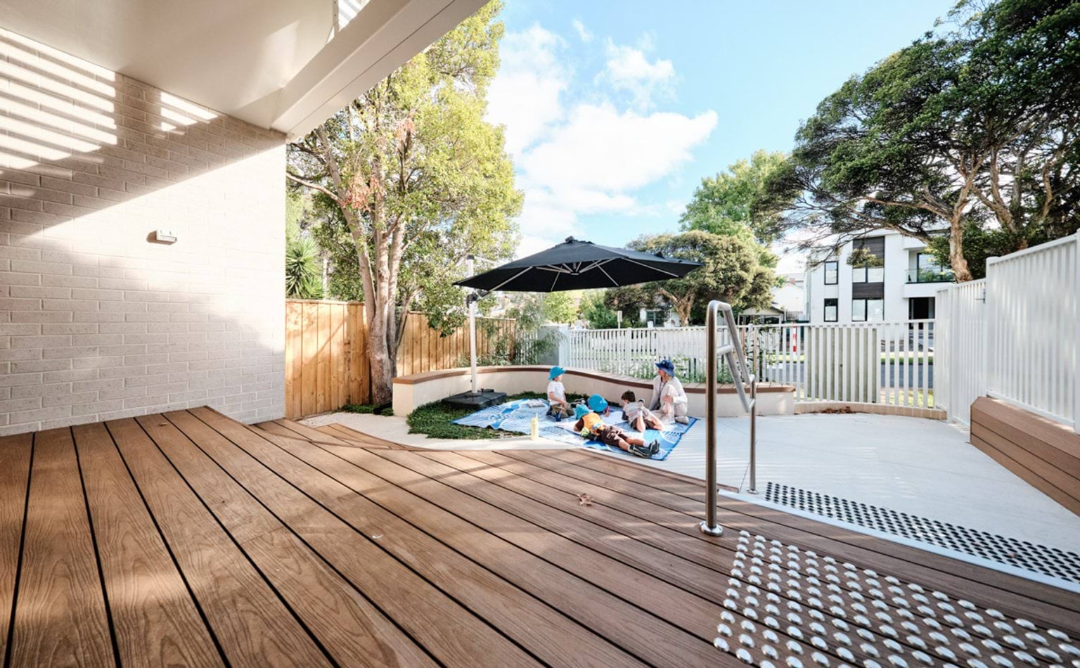 An outdoor play area with timber steps leading to glass doors. A curved seating wall surrounds a garden bed, and a large black umbrella shades a blue picnic rug.
