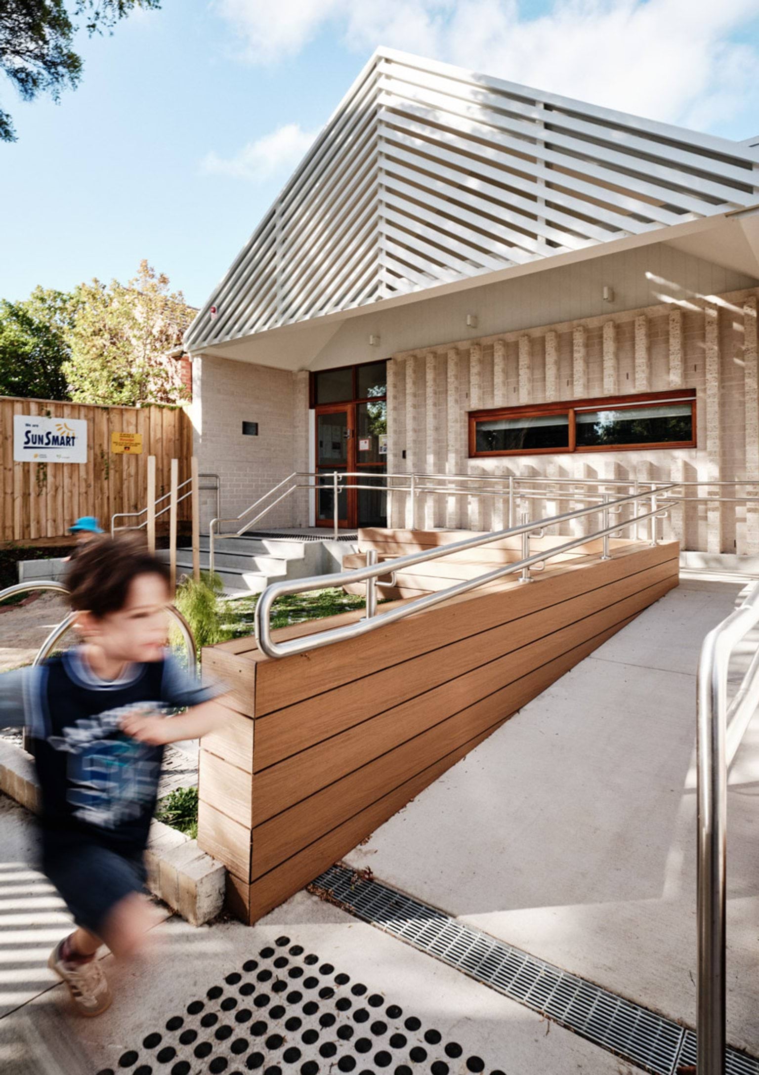 The entrance of the kindergarten with cream brick walls, a slatted white roof feature and timber-framed windows. A ramp with stainless steel handrails leads to the doorway, and a SunSmart sign is on the fence.